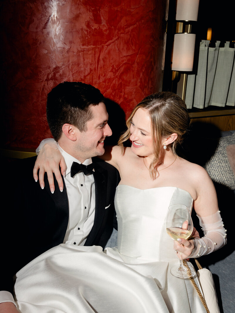 Direct flash photo of bride and groom cuddling in a hotel bar, smiling at each other. The bride has a glass of wine in her hand.