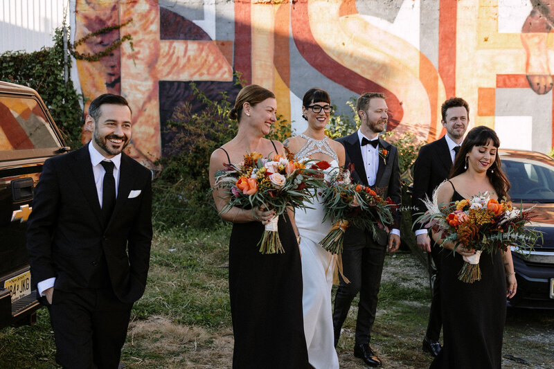 A wedding party walks together, wearing elegant black attire and carrying colorful bouquets, with a vibrant mural in the background.