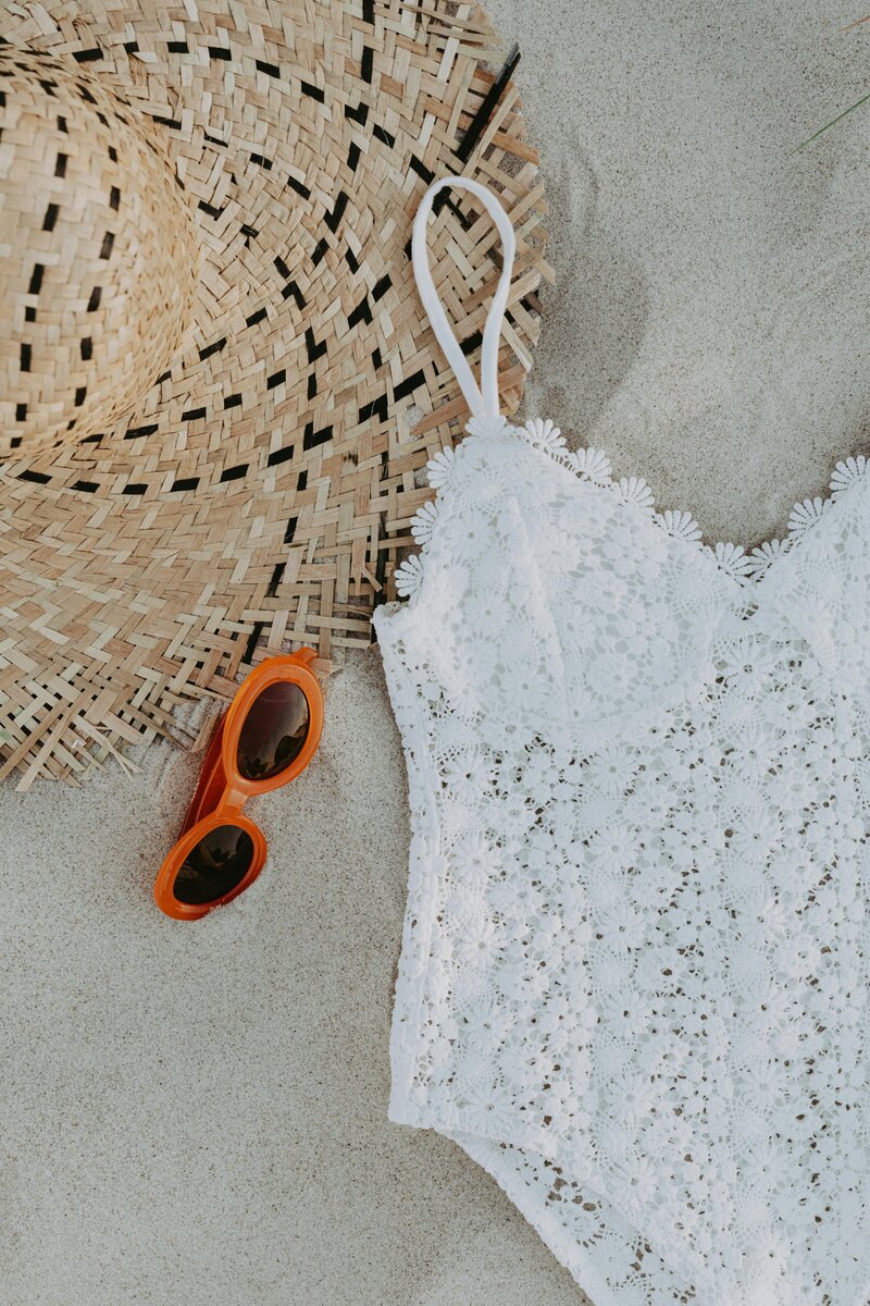 white swimsuit, straw hat, and orange sunglasses laying in the sand