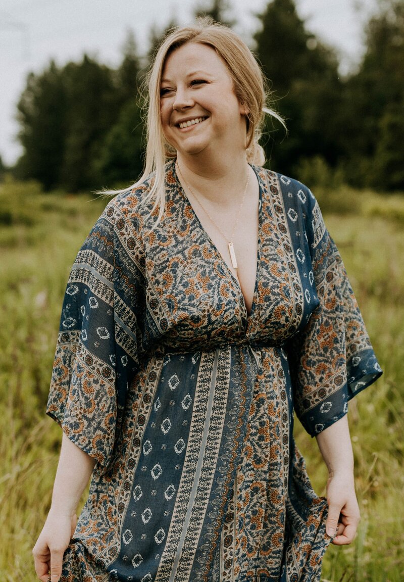 Jaymie Sutherland, Spiritual Healer, Activator, & Expander, standing with a bridge in the background, looking over to the left with a hands clasped in front of her & a soft smile on her face.