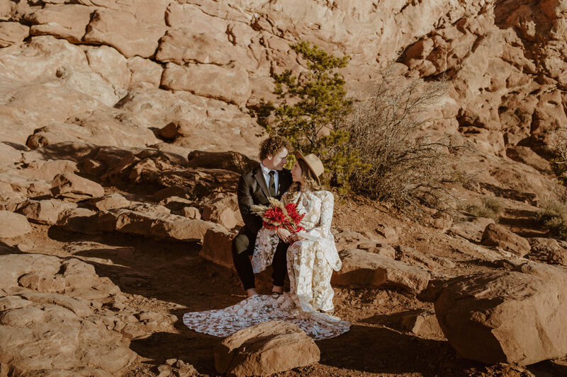 Bride wearing white boho dress holding red bouquet sitting next to the groom wearing a black suit on the red rocks at Arches National Park.
