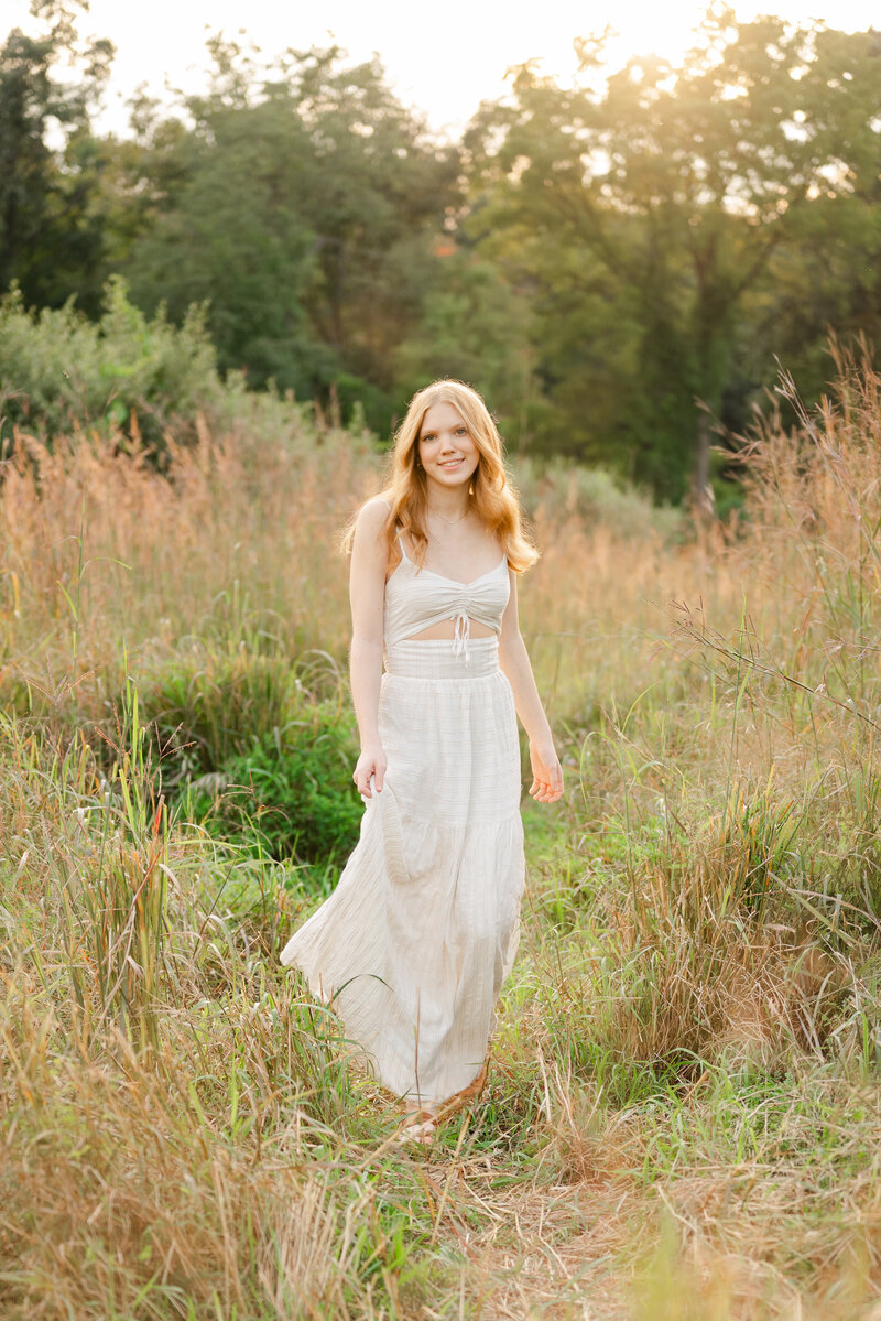 Senior girl wearing a white dress, standing in an autumn field with colorful foliage.
