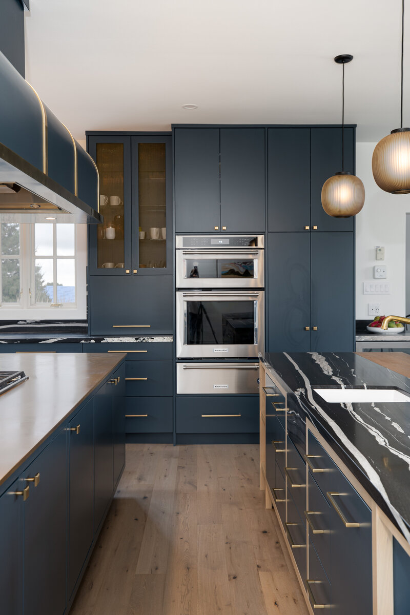 A brass and a stone kitchen island framed by deep blue cabinetry, sleek silver ovens, and moody hanging pendant lights