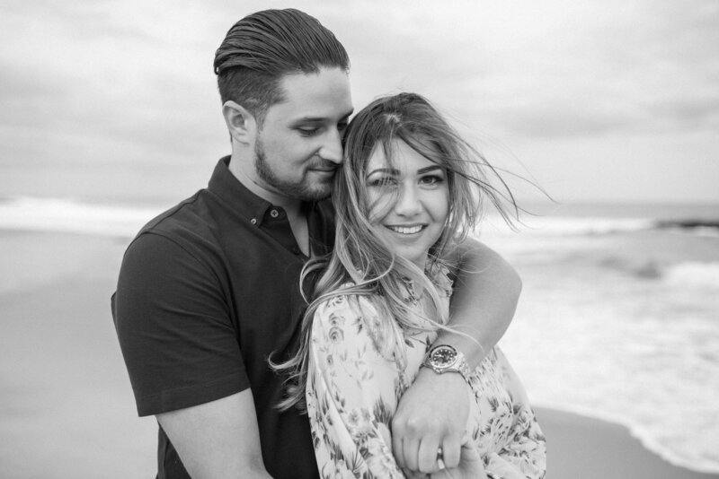A couple embraces on a beach, with waves gently lapping at the shore, captured in a romantic black-and-white photo.