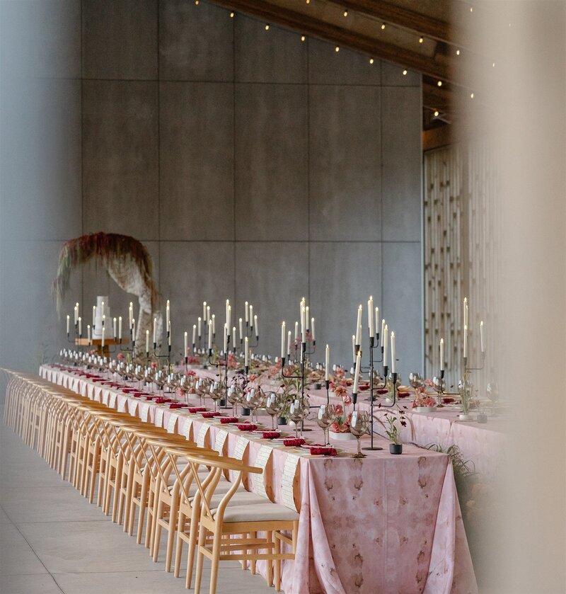 Editorial wedding reception at Gather Greene in the Catskills, New York, featuring a long banquet table with blush-pink linens, tall black taper candles, and minimalist floral arrangements beneath a wooden pavilion with concrete walls — a modern Catskills wedding blending sculptural design and romantic minimalism.
