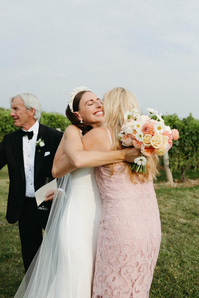A bride in a white dress and veil hugs a woman in a pink dress, holding a bouquet of flowers in an outdoor setting. A man in a black suit stands nearby, all captured beautifully by a renowned Muskoka wedding photographer.