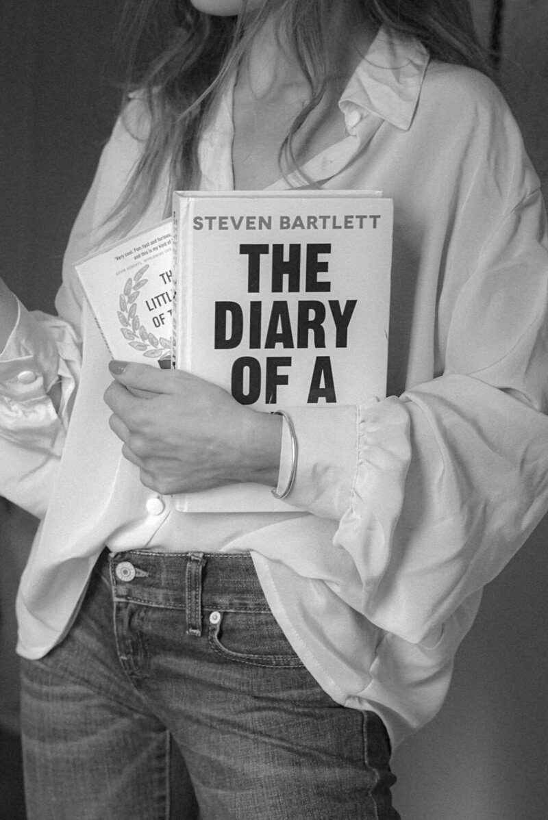 A woman with long blonde hair, wearing an oversized cream silk blouse and jeans, holds a stack of books close to her chest, exuding understated sophistication.