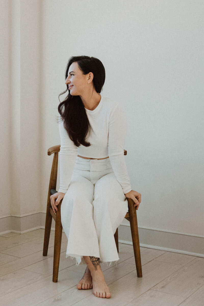 Holistic Esthetician Jen Stoeckert of Minimal Beauty sitting in a wooden chair looking to the side wearing a white long sleeve top and white pants.