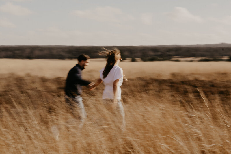 Een man en vrouw rennen samen door een uitgestrekt veld, omringd door groene gras en bloemen.