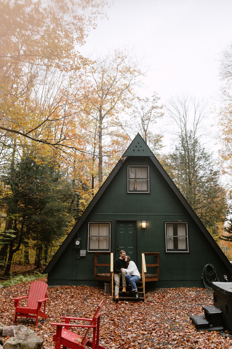 A couple sits cozily on the porch of a green cabin, surrounded by nature, sharing a peaceful moment together.