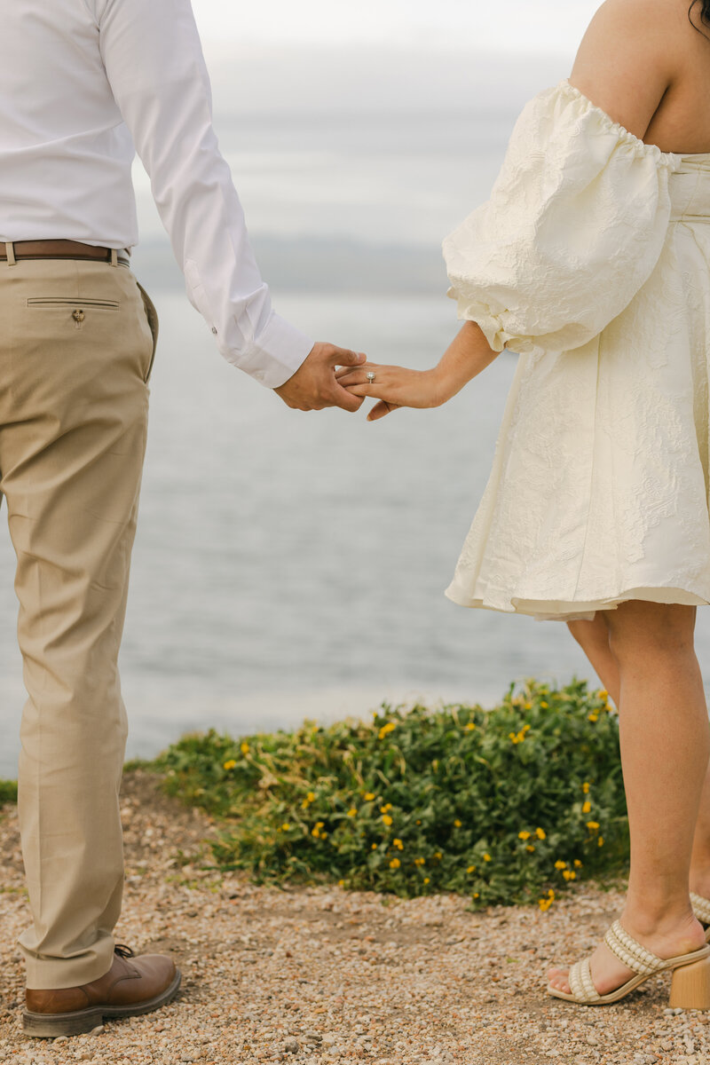 Editorial close-up of a couple holding hands on the beach, featuring cream-toned fashion styling, heels in the sand, and a soft, romantic atmosphere — perfect for luxury wedding and engagement photography in Monterey California.