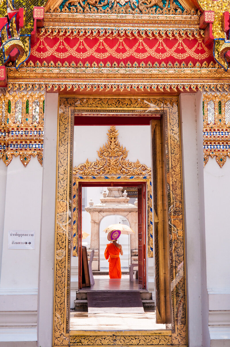 Person in orange robes holding a parasol, framed by ornate golden temple doorways in a traditional Asian temple.