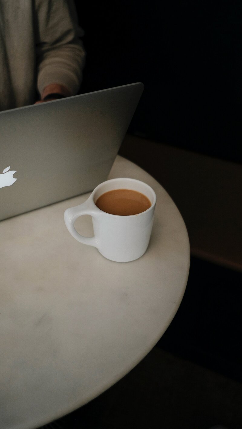 a milky coffee in a white mug sits on a table near a silver macbook