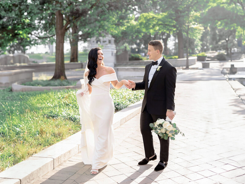 A bride in an off-shoulder gown and a groom in a tuxedo walk hand in hand along a sunlit, tree-lined path. The bride holds her dress while the groom carries a bouquet of white roses. Both are smiling and gazing at each other.