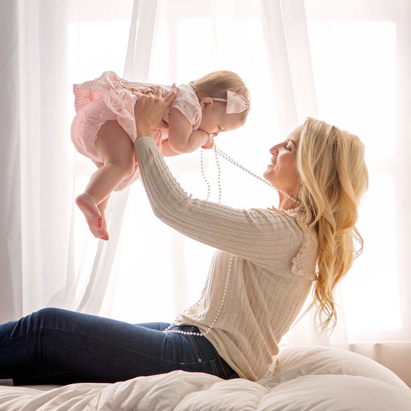 Sky 9 Studio | Young blonde mother holding baby girl in the air in front of large bright and airy window while she puts her mother's pearls in her mouth