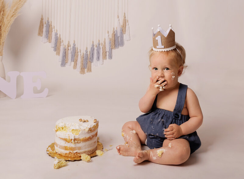 Little boy wearing denim overalls and a first birthday crown smiles at the camera as he eats cake for his first birthday cake smash photoshoot in Brooklyn, NY. Captured by premier Brooklyn NY family photographer Chaya Bornstein Photography.