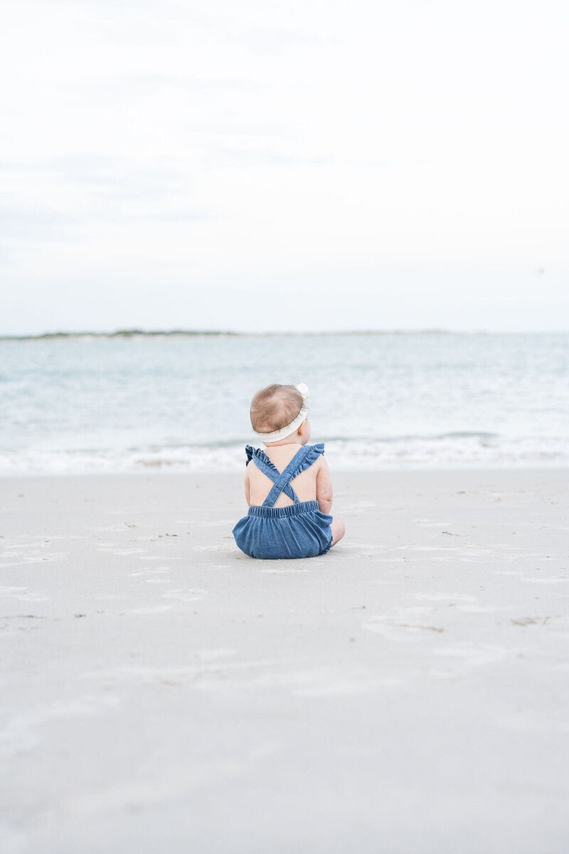 baby girl wears denim overalls sitting on the beach with her back to the camera