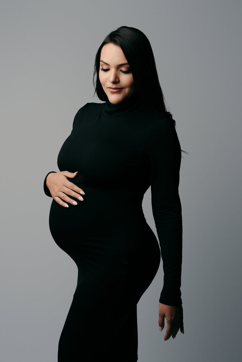 Pregnant woman looking at camera wearing black maternity dress and posing holding baby bump
