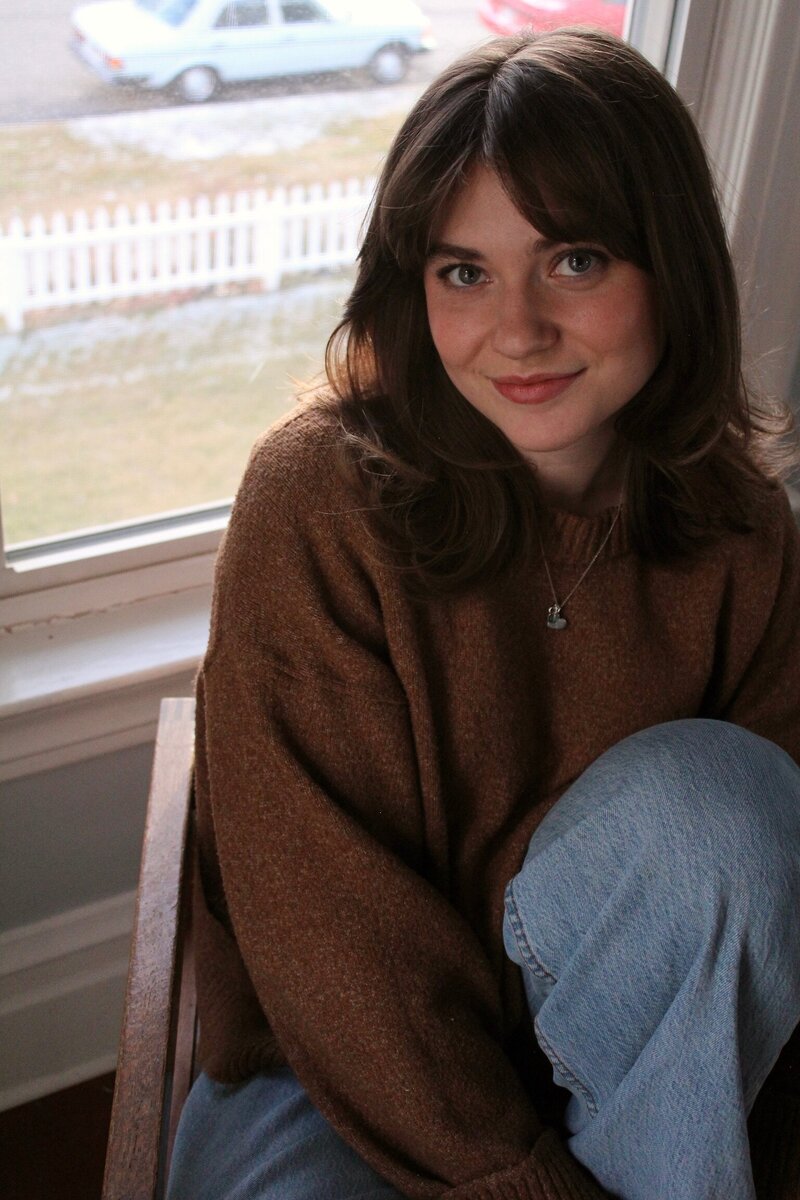 Katie Graper, Showit web designer, sitting in a char and softly smiling up at the camera with one foot propped on the seat