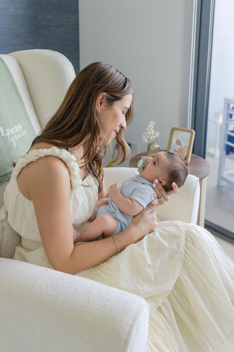 A mother in a cream dress lovingly gazes at her baby boy while holding him in a glider chair. The baby wears a blue outfit with a pacifier, and the nursery features a framed photo, vase of flowers, and soft natural light from a nearby window.