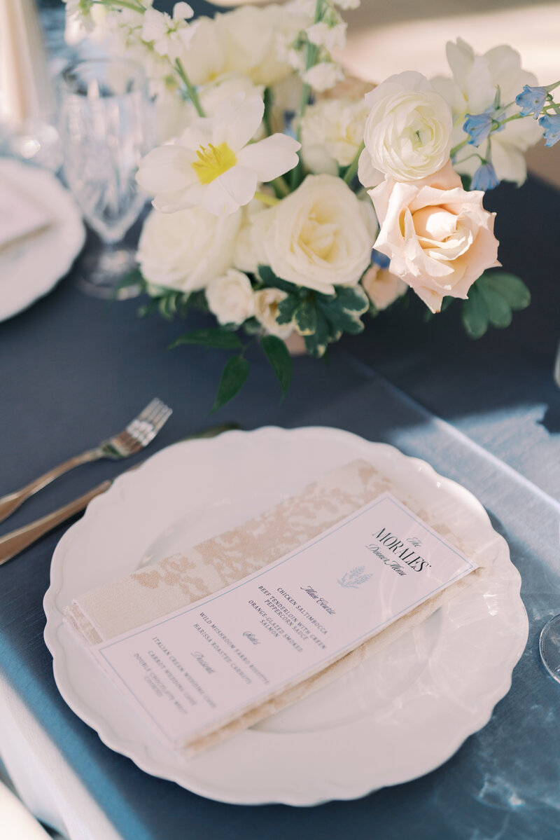 Elegant wedding place setting with custom printed menu, neutral napkin, and white floral centerpiece on a navy linen. 