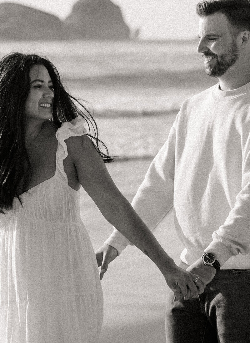 Black and white photo of a couple holding hands on Cannon Beach with sea stacks in the background.
