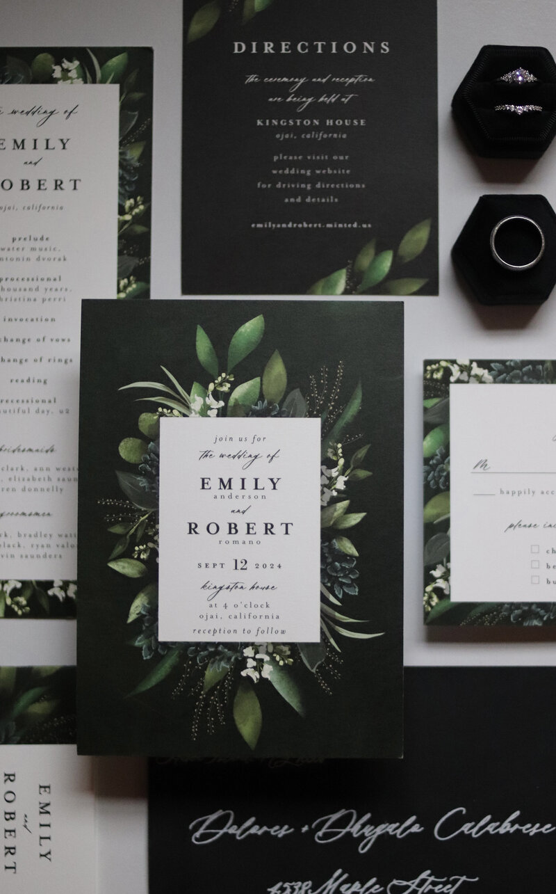 A selection of black and green wedding invitations and enclosurres are displayed with an envelope wedding program velvet ring box and wedding and engagement rings