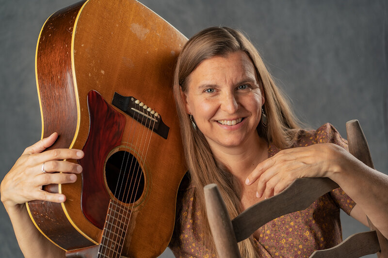 Woman with guitar on her shoulder, smiling, in a brown dress. 