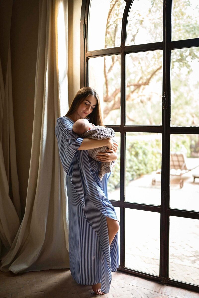 mère en robe bleur avec son bébé dans les bras devant une grande fenêtre