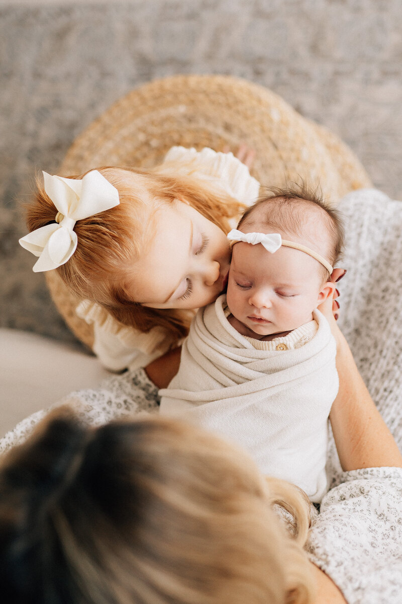 view from above of mother holding swaddled newborn while big sister kisses her cheek