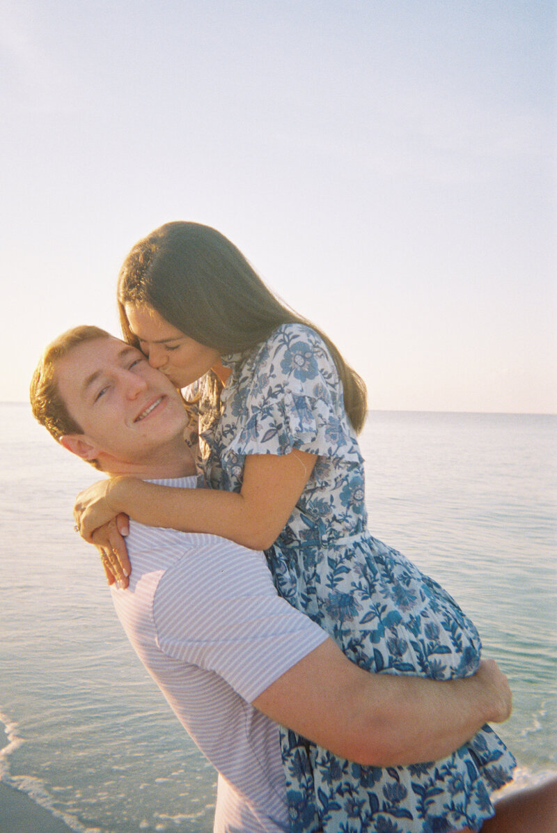 Man holding woman while she kisses him on the cheek with ocean in the background