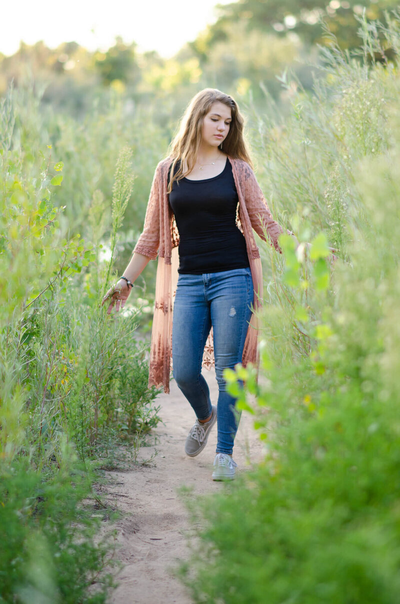 girl in a pink sweater walking through some tall plants on a path and touching the plants