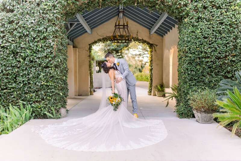 Groom dips bride in an embrace in front of arched garden entrance