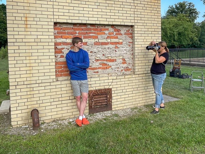 female photographer taking picture of a high school senior boy in a blue sweatshirt and gray shorts.  The boy is standing with arms crossed against a beige brick wall