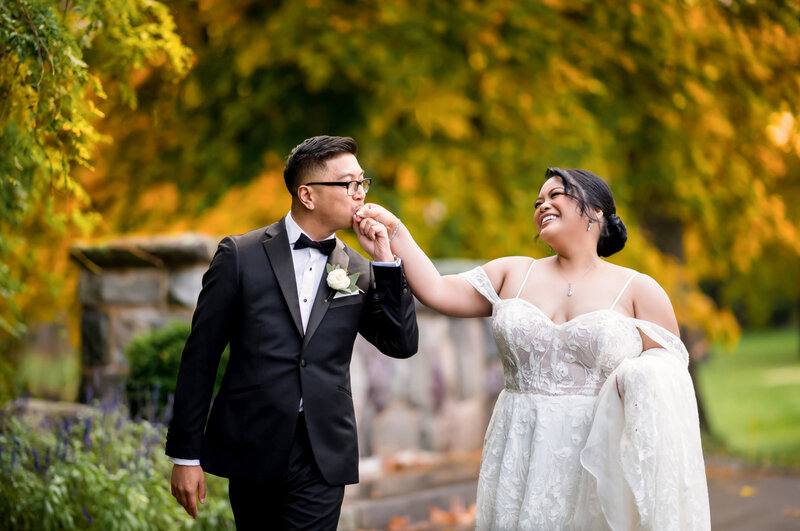 Groom kissing his bride's hand on the grounds of Brooklake Country Club in Florham Park NJ with fall foliage behind them