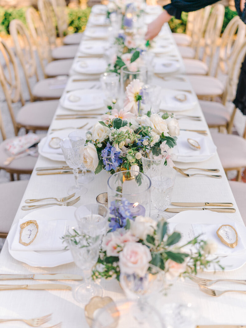 Long outdoor wedding table decorated with white linens, gold cutlery, vintage plates, and pastel floral centerpieces.