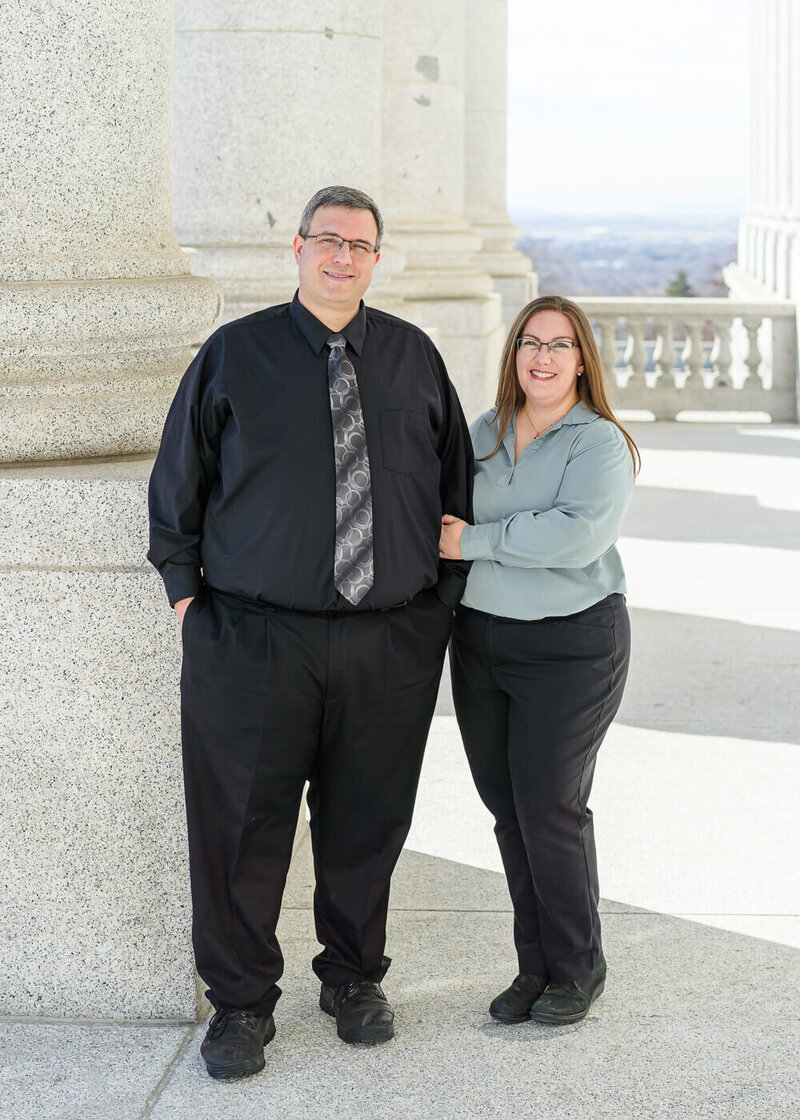 a husband wearing a black dress shirt, black slacks, and a silver tie stands next to his wife wearing a sea foam green blouse and black slacks  stand outside the utah state capitol