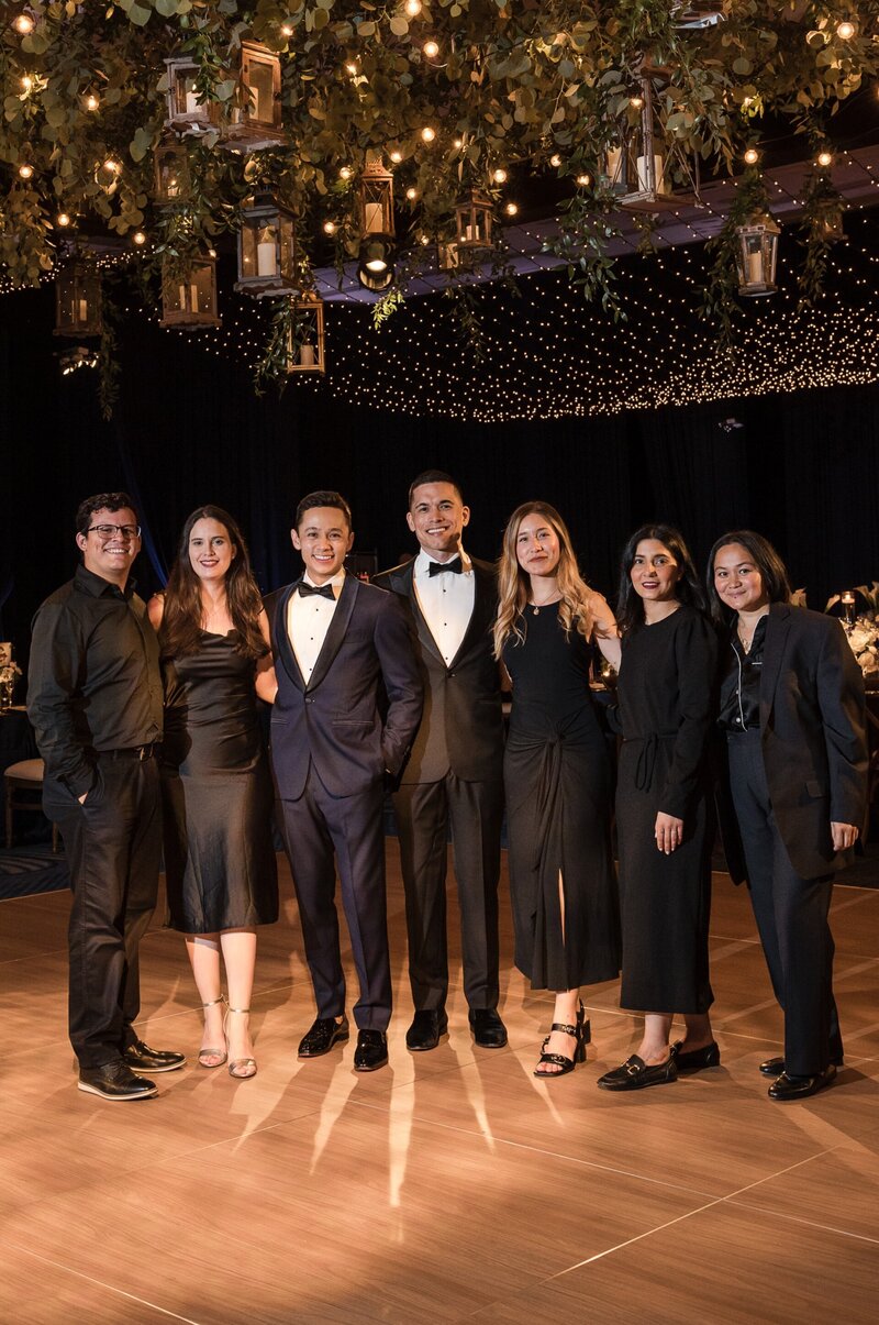 A group of seven people in formal attire stand under a decorative ceiling with lights and foliage.