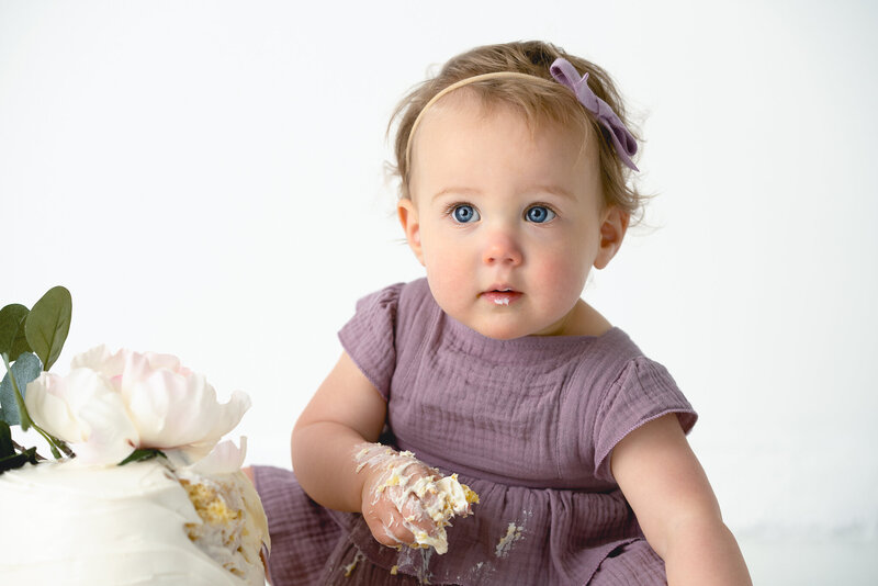 A baby girl in a purple outfit is sitting in front of a birthday cake for her smash cake session.