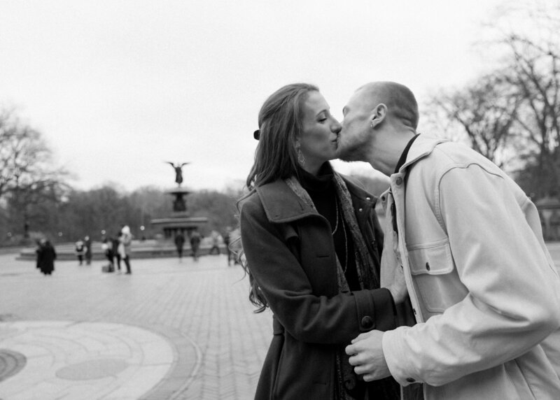 Black and white image of couple kissing outside with people and a fountain in the background
