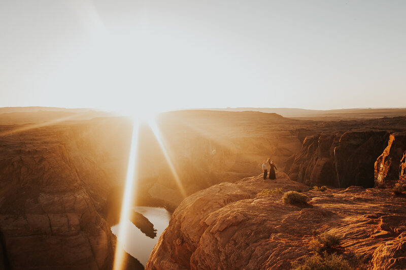 A couple stands together on a cliff's edge, looking out over a stunning canyon landscape as the sun sets, casting a warm glow over the scene in Page, Arizona.