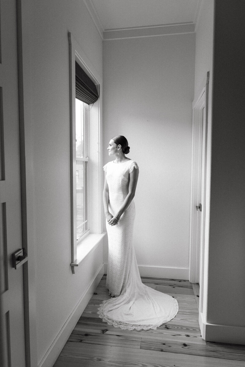 Black and white bridal portrait of bride standing by window in soft morning light, captured in timeless documentary style.