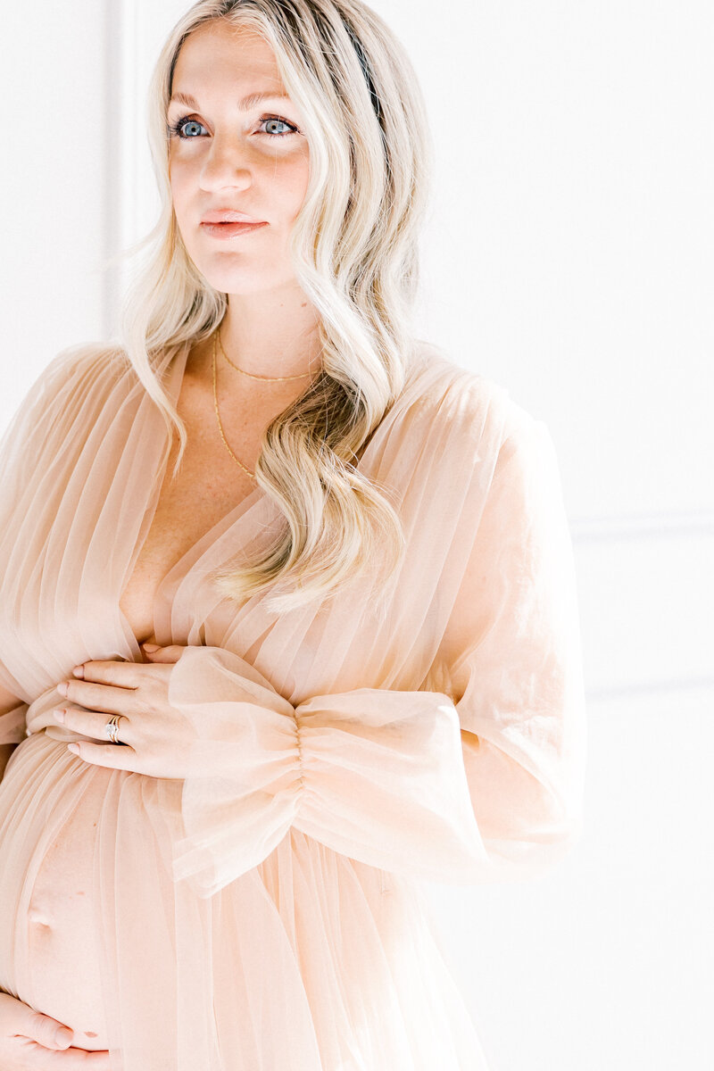 A blonde mother to be stands in a window holding her bump in a pink maternity gown supplied by a Charlotte Family Photographer