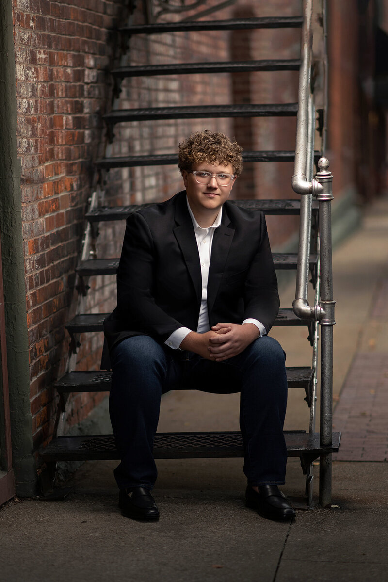 A high school senior sits in a sports coat on alley fire escape stairs