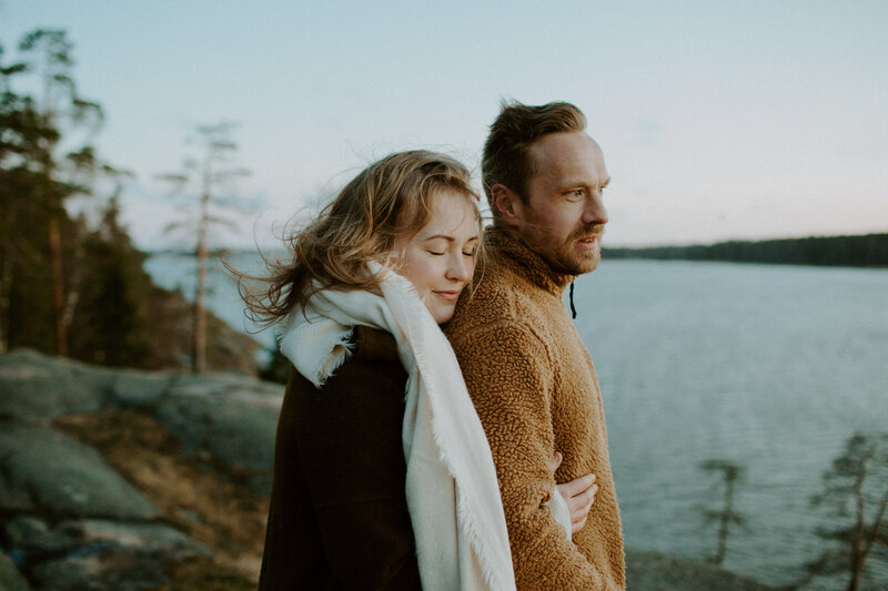Pari seisoo rantakalliolla nainen halaten miestä selän takaa ja nojaten päätä miehen olkapäähän silmät suljettuina Kivenlahdessa Espoossa