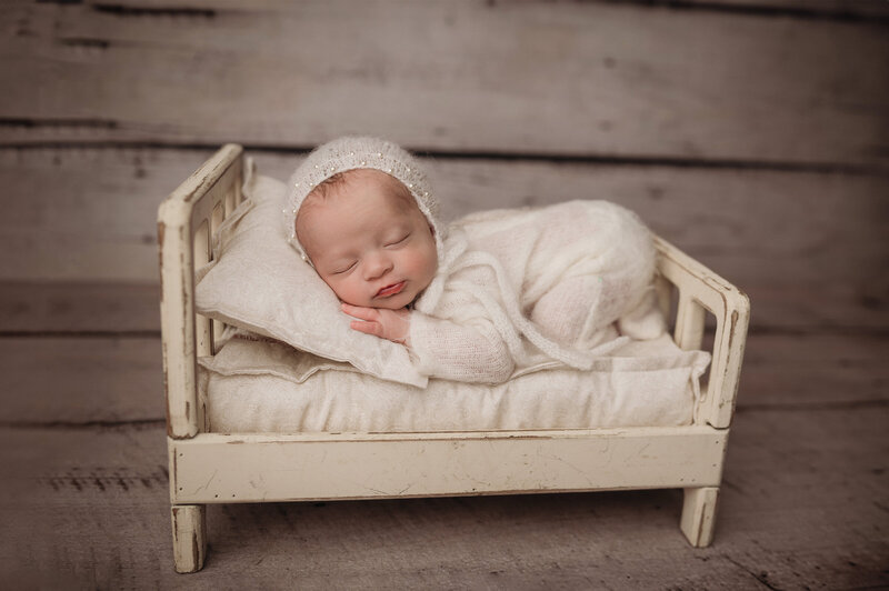 Ct  newborn photographer captures two week old sleeping baby girl wearing a  fuzzy white onesie with a matching bonnet laying in a little white bed prop on her belly  in front of white washed wood backdrop