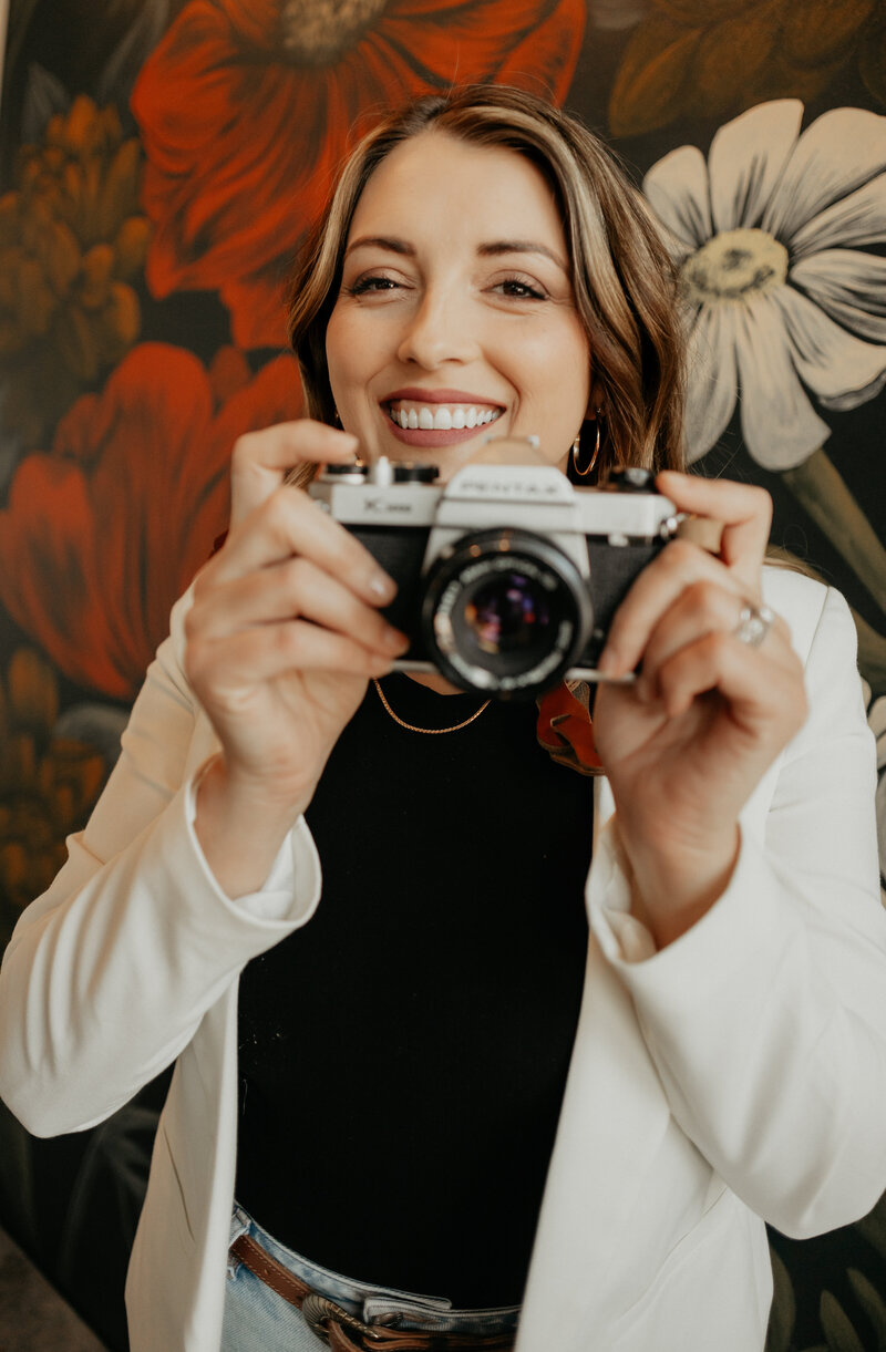 A photo of Brooke Ward holding a camera and smiling in front of a wall with floral wallpaper.