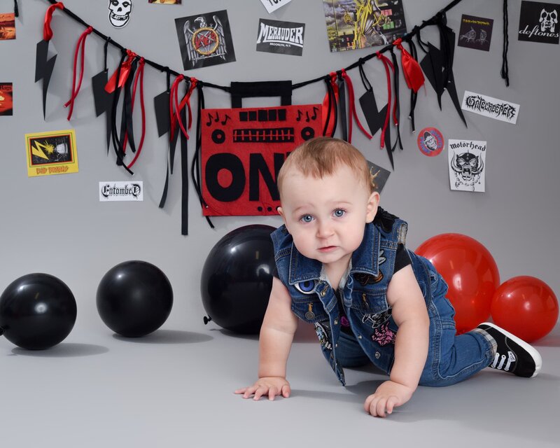 A baby boy dressed in a denim vest and jeans crawling on the floor with a rockstar-themed backdrop featuring black and red balloons and a "ONE" sign.