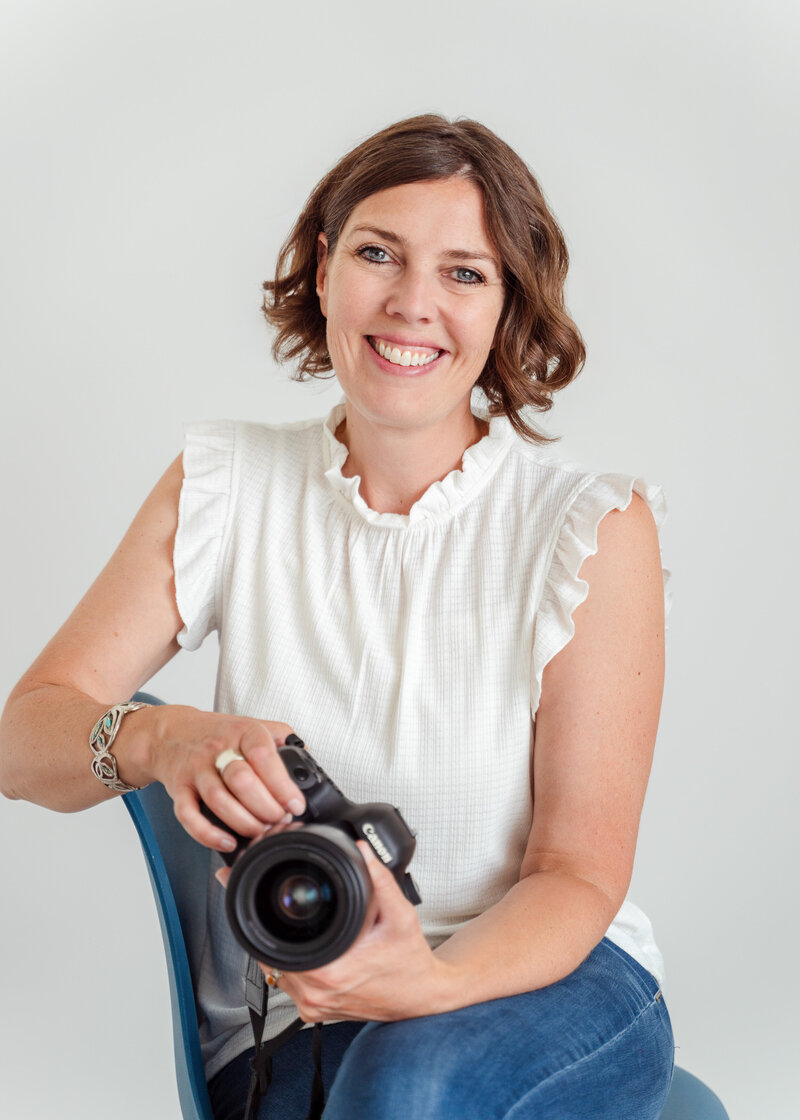 Calgary Brand Photographer Kate Daniel Photography, smiling, wearing a white shirt and blue jeans, sitting on a chair and holding a camera.