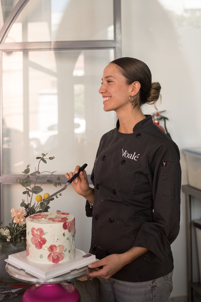 A smiling pastry chef from Voalé Bakery stands beside a floral-themed cake in a minimalist kitchen space.